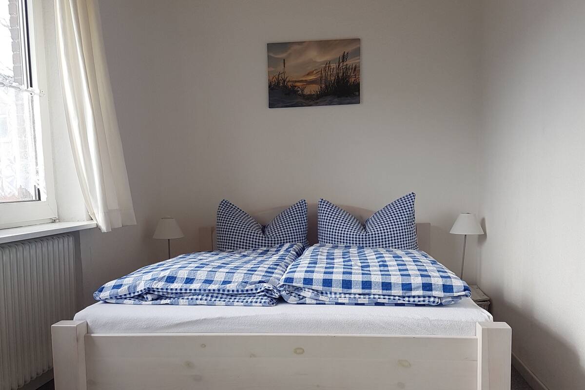 Un dormitorio con cama blanca, ropa de cama a cuadros, un cuadro en la pared y una ventana con cortinas.