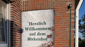 En byggnad av tegel med en skylt som lyder: ”Hjärtligt välkommen till Birkenhof” och en blomsterrabatt framför.