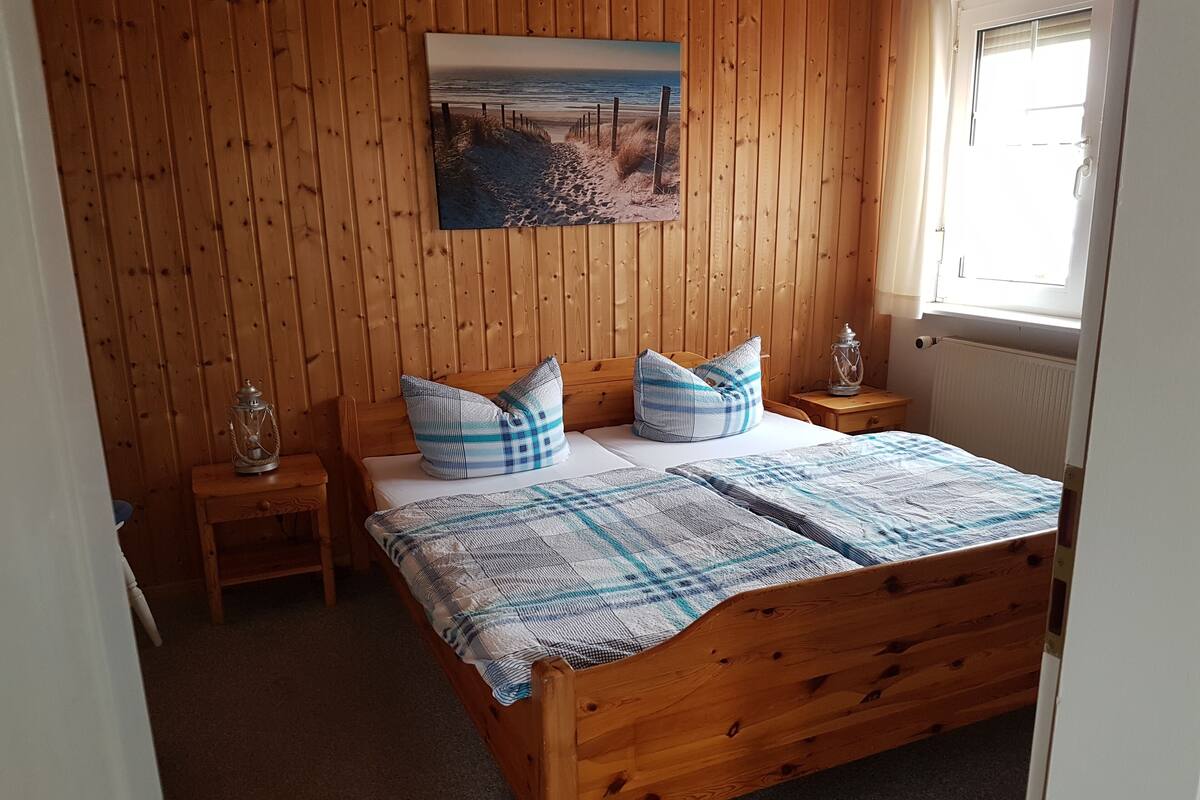 Dormitorio con paneles de madera, una cama, dos mesitas de noche, un cuadro en la pared y una ventana.