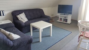 Ett vardagsrum med en mörkblå soffa, ett vitt soffbord, en platt-TV på ett vitt TV-bord och ett litet vitt sidobord med en lampa.