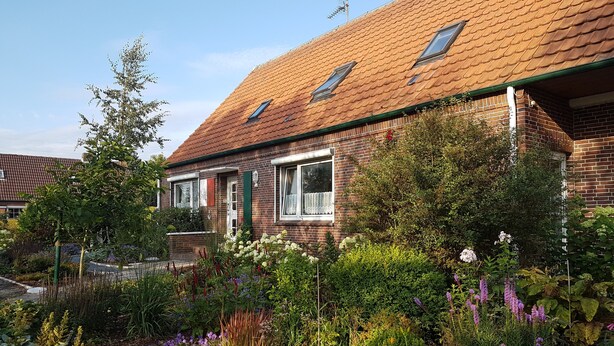 Ett hus byggt av tegel med ett tak täckt av takpannor, flera fönster och en trädgård med olika växter och blommor.