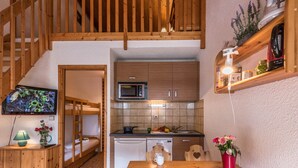Eine kompakte Küche mit Holdekor-Schränken, einer Mikrowelle und einem Spülbecken. Ein Holzesstisch mit Stühlen und ein an der Wand montierter Fernseher sind ebenfalls vorhanden.