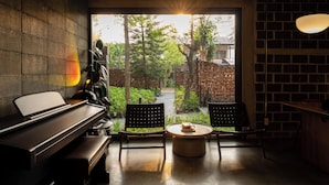 Une pièce avec un piano, deux chaises, une table ronde et une vue sur un jardin, visible à travers une grande fenêtre.
