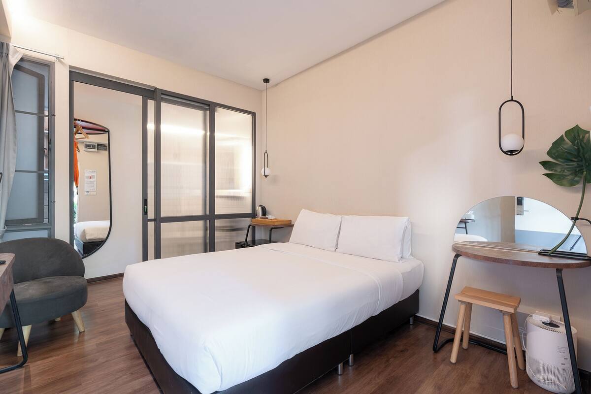 A modern hotel room with a large bed, a desk, a chair, a small stool, a mirror, and a potted plant.