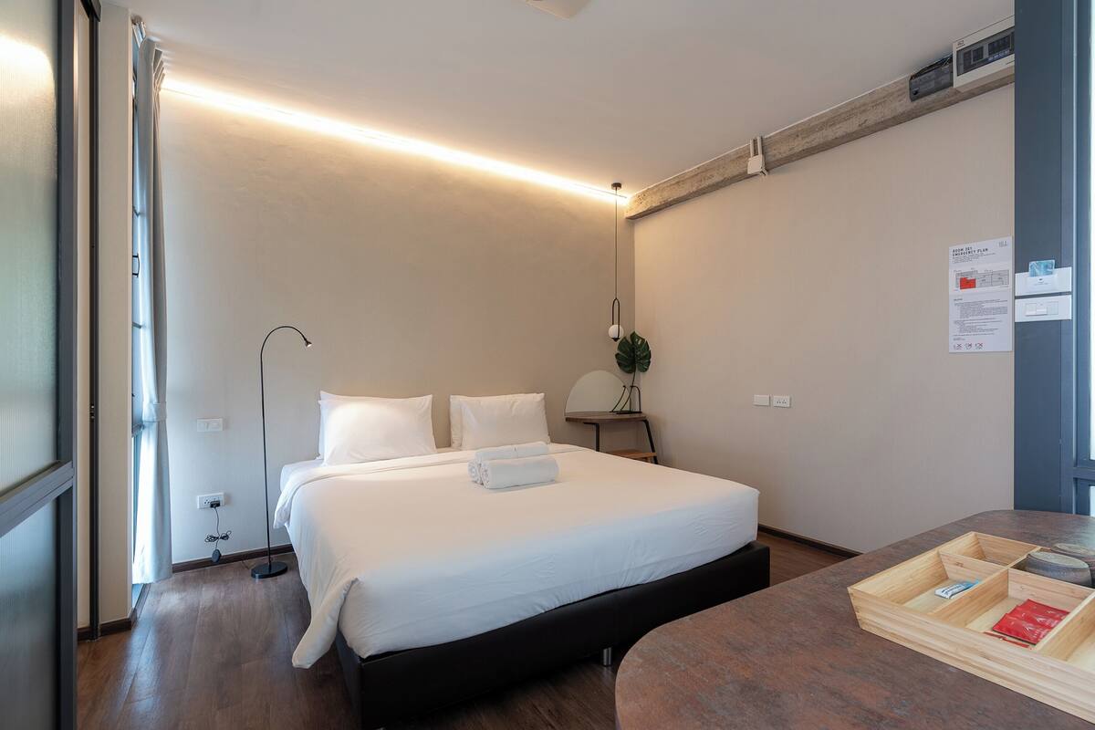 A modern hotel room with a large bed, bedside table, and a wooden side table with a lamp.