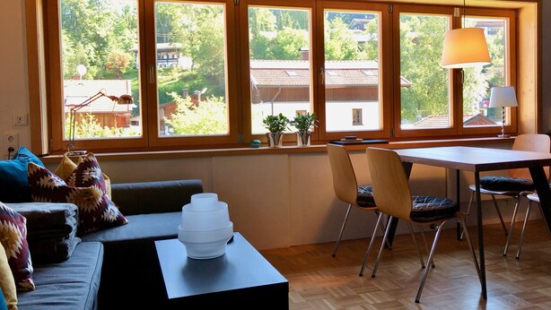 A room with a large window, a dining table, chairs, a sofa, and a lamp. - Apartment "Am Berg" in Fischbachau, Birkenstein (Fischbachau)