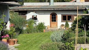 A house with a white exterior, a wooden door, and a small garden with potted plants and a lawn. - Apartment "Am Berg" in Fischbachau, Birkenstein (Fischbachau)
