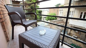 Un balcón con una mesa y silla de mimbre, un cenicero de vidrio transparente y vistas de los edificios circundantes.