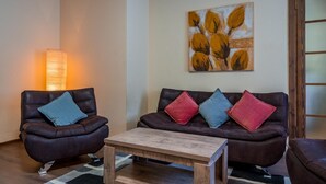 Una sala de estar con dos sillones de cuero, una mesa de centro de madera y un sofá con cojines de colores. Un cuadro cuelga en la pared, y una lámpara está a la izquierda.