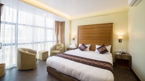 Hotelkamer met een groot bed, een houten hoofdbord, een zithoek met een fauteuil, een nachtkastje en een groot raam met gordijnen.