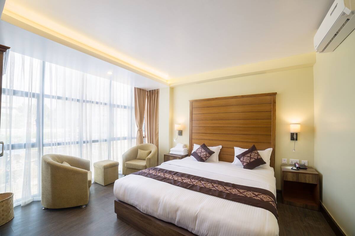Una habitación de hotel con una cama grande, cabecera de madera, zona de estar con una silla, mesita de noche y un ventanal con cortinas.