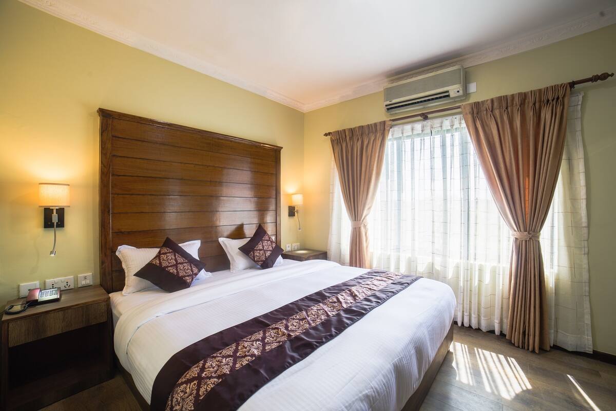 Habitación de hotel con una cama grande, cabecera de madera, dos mesitas de noche con lámparas, un teléfono y una ventana con cortinas.