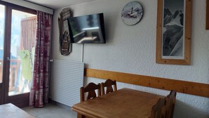 A room with a wooden dining table, chairs, a television mounted on the wall, a clock, and a framed picture.