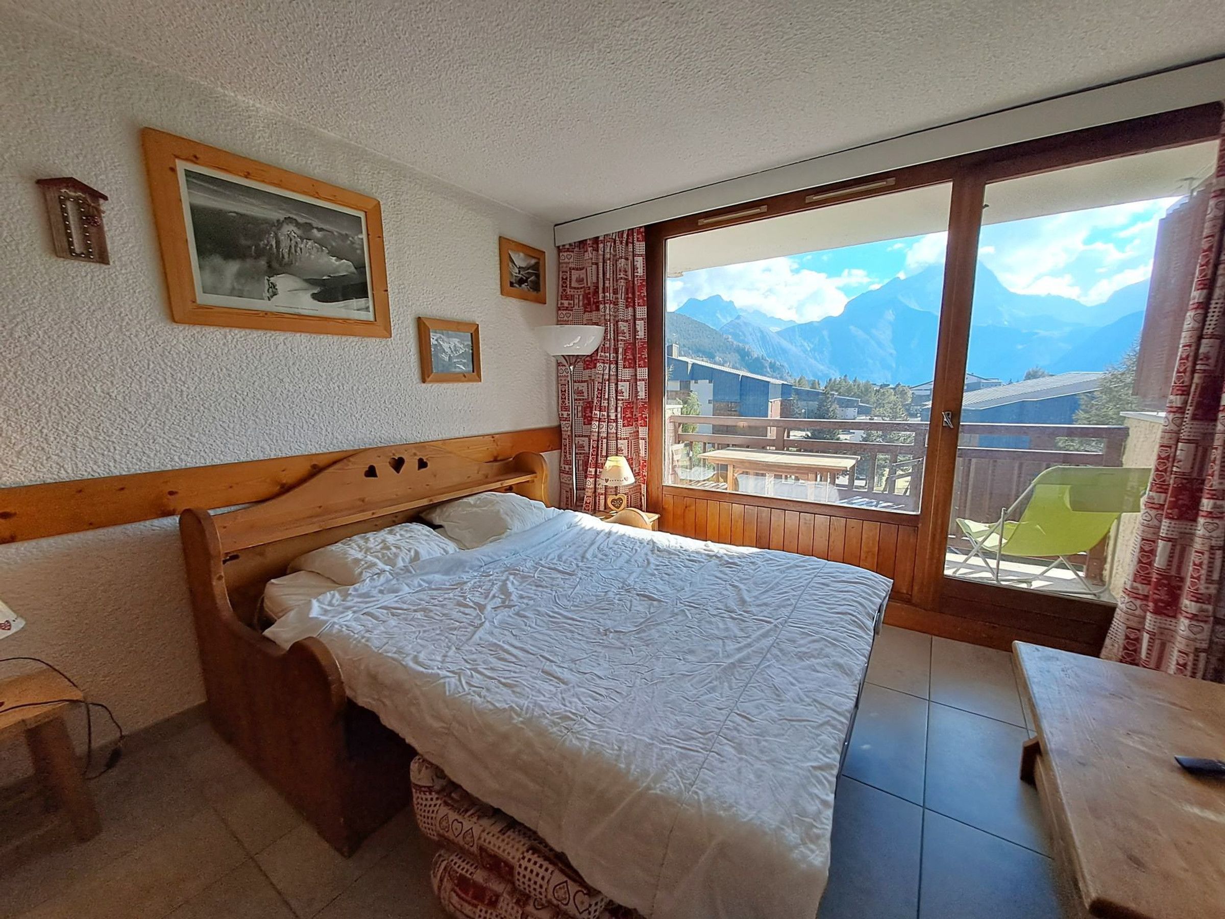 A bedroom with a wooden headboard, a bed with white bedding, a small bedside table, a window with a view of mountains, and a balcony with a chair.
