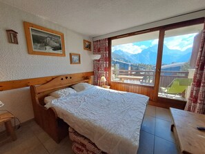 A bedroom with a wooden headboard, a bed with white bedding, a small bedside table, a window with a view of mountains, and a balcony with a chair.
