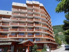 A multi-story hotel with balconies, a ski resort in the background, and a clear blue sky.