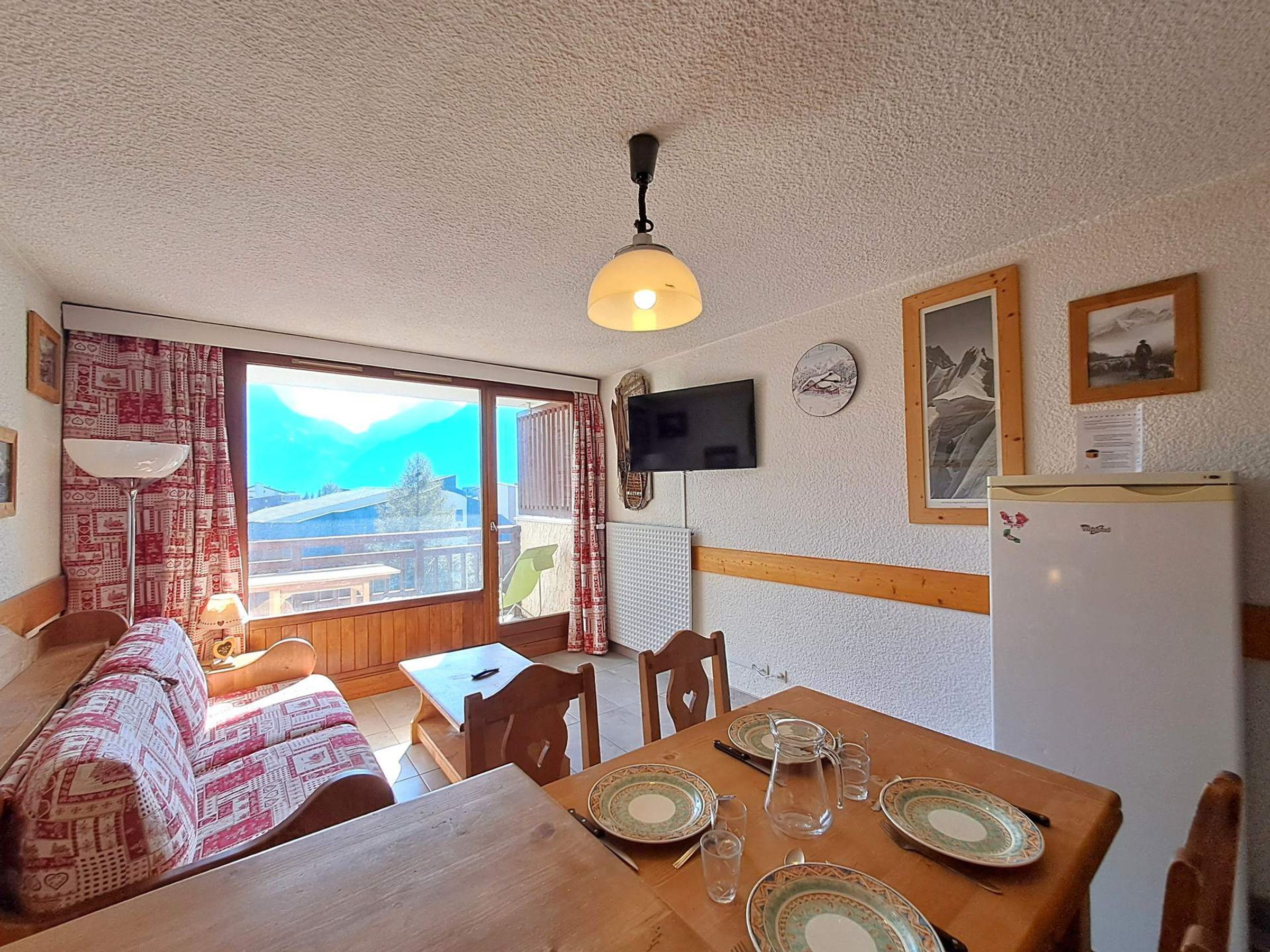 Een eethoek met een tafel gedekt voor twee personen, een aan de muur bevestigde televisie en een uitzicht op bergen door het raam.