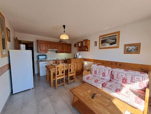 Une cuisine de taille réduite avec des meubles en bois, un réfrigérateur blanc, une table à manger avec des chaises et un canapé recouvert d’un tissu à motifs.