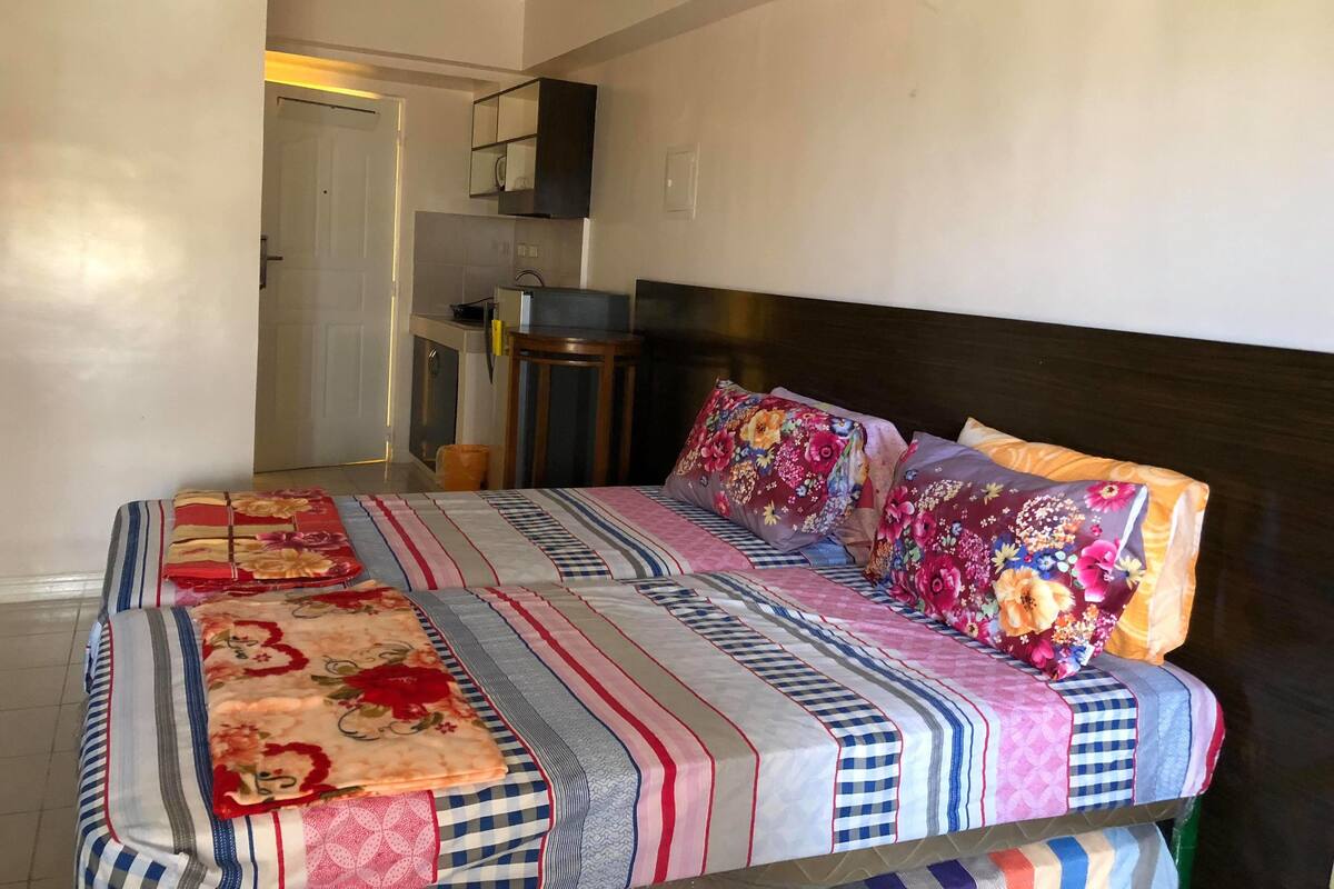 Quarto de hotel com uma cama grande, uma mesa pequena e uma kitchenette ao fundo.