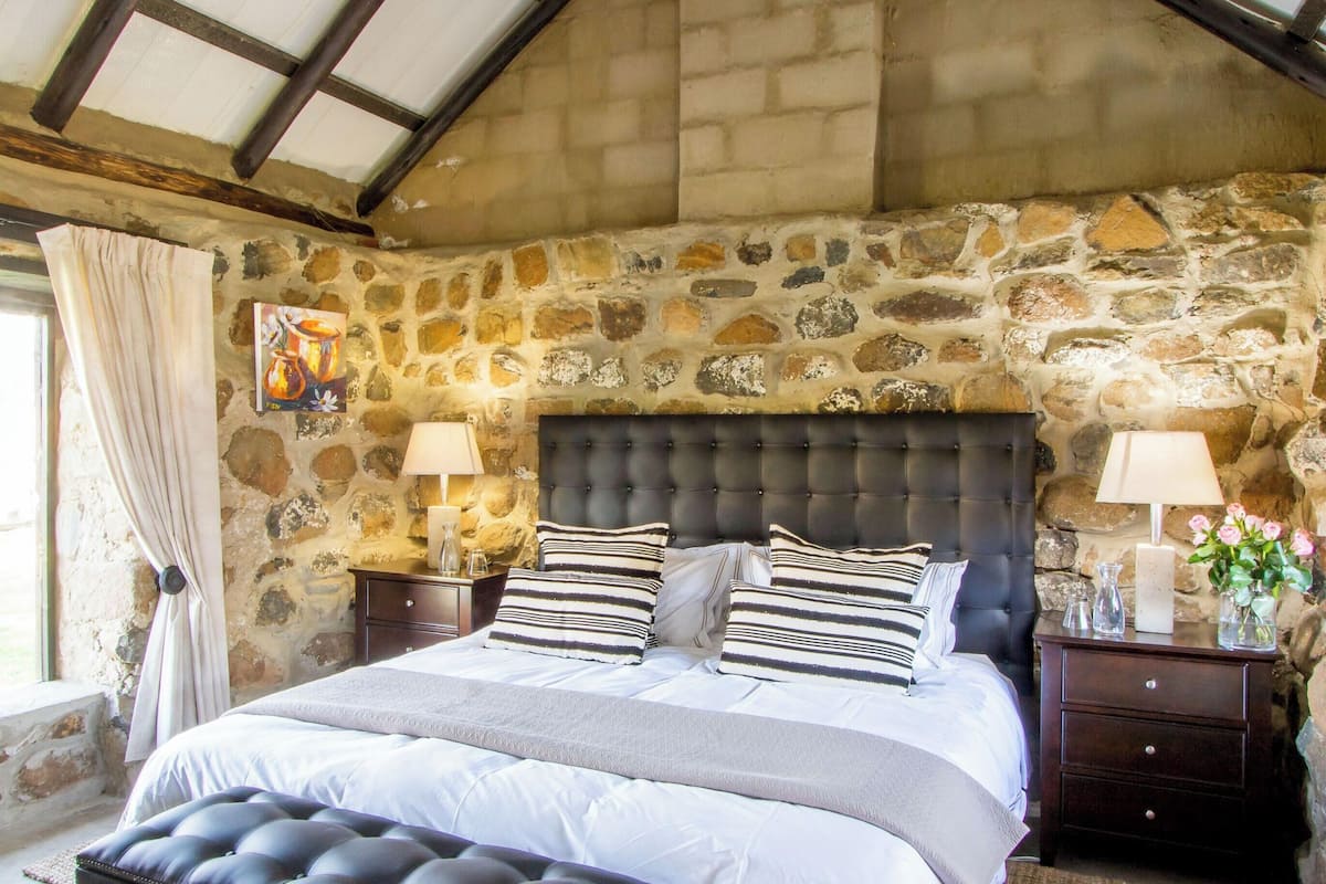 A bedroom with a stone wall, a large bed with striped pillows, two bedside tables with lamps, and a window with curtains.