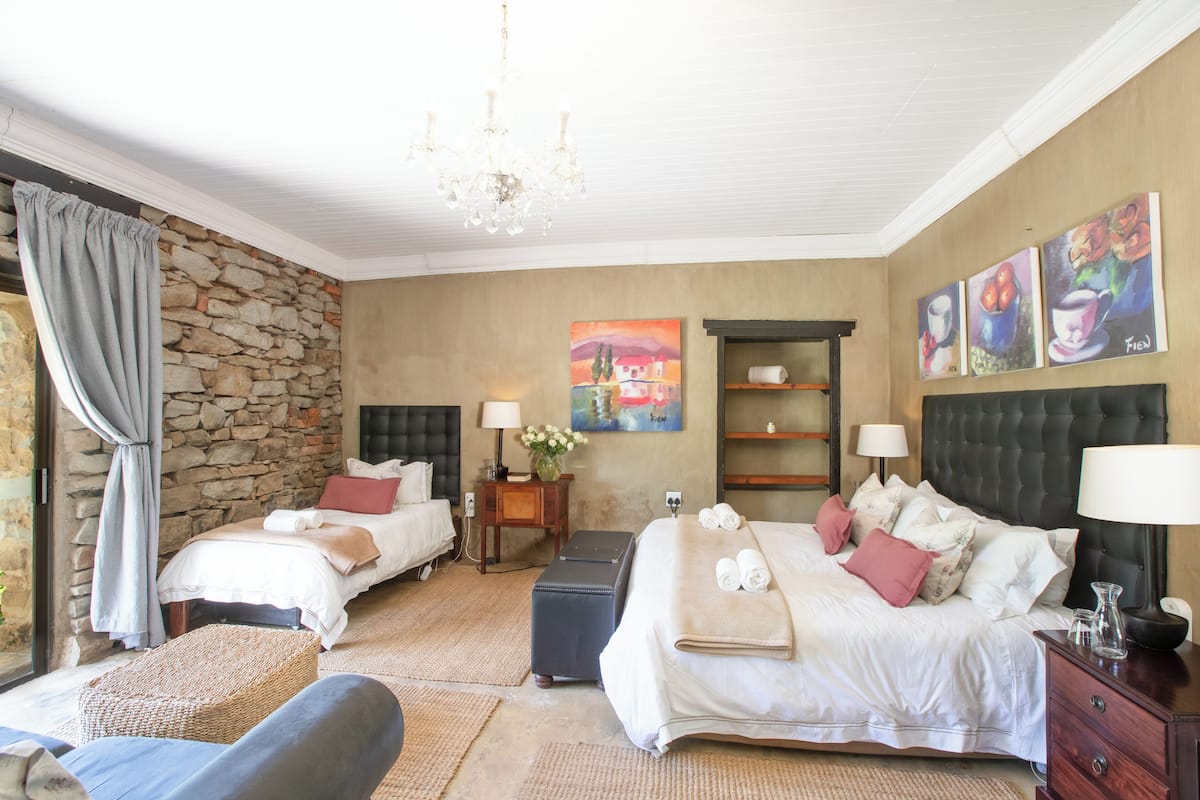 A bedroom with two beds, a stone wall, a chandelier, and artwork on the wall.