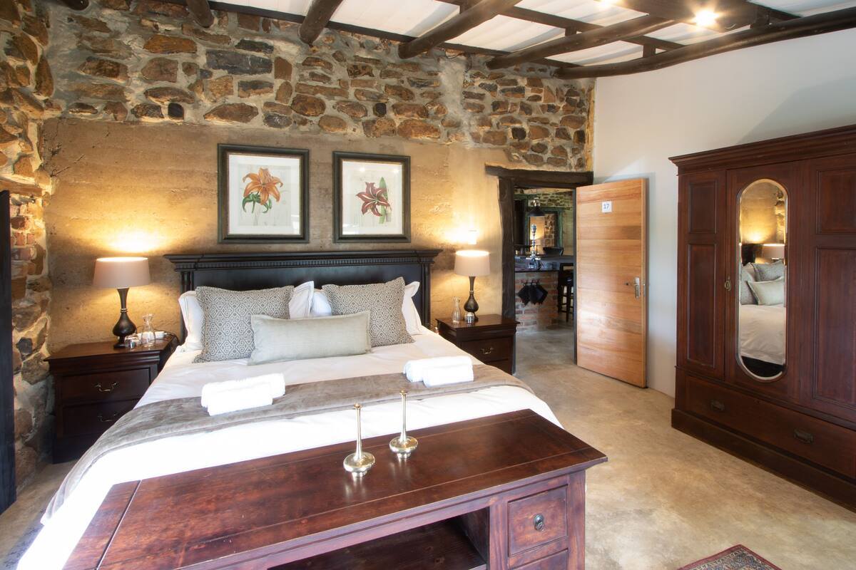 A bedroom with a stone wall, a wooden bed with a headboard, two bedside tables with lamps, a large wooden wardrobe, and a mirror.
