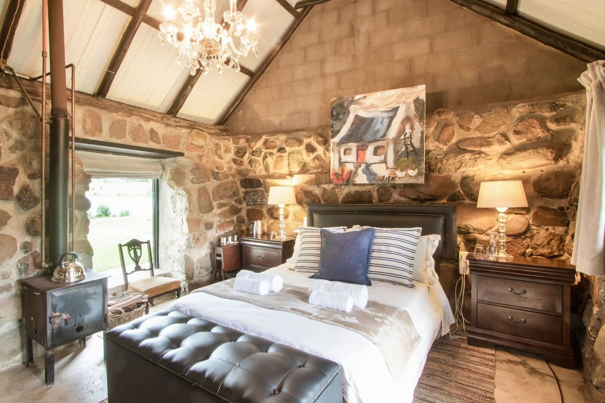 A bedroom with a stone wall, a bed with a headboard, bedside tables, a chandelier, and a painting.