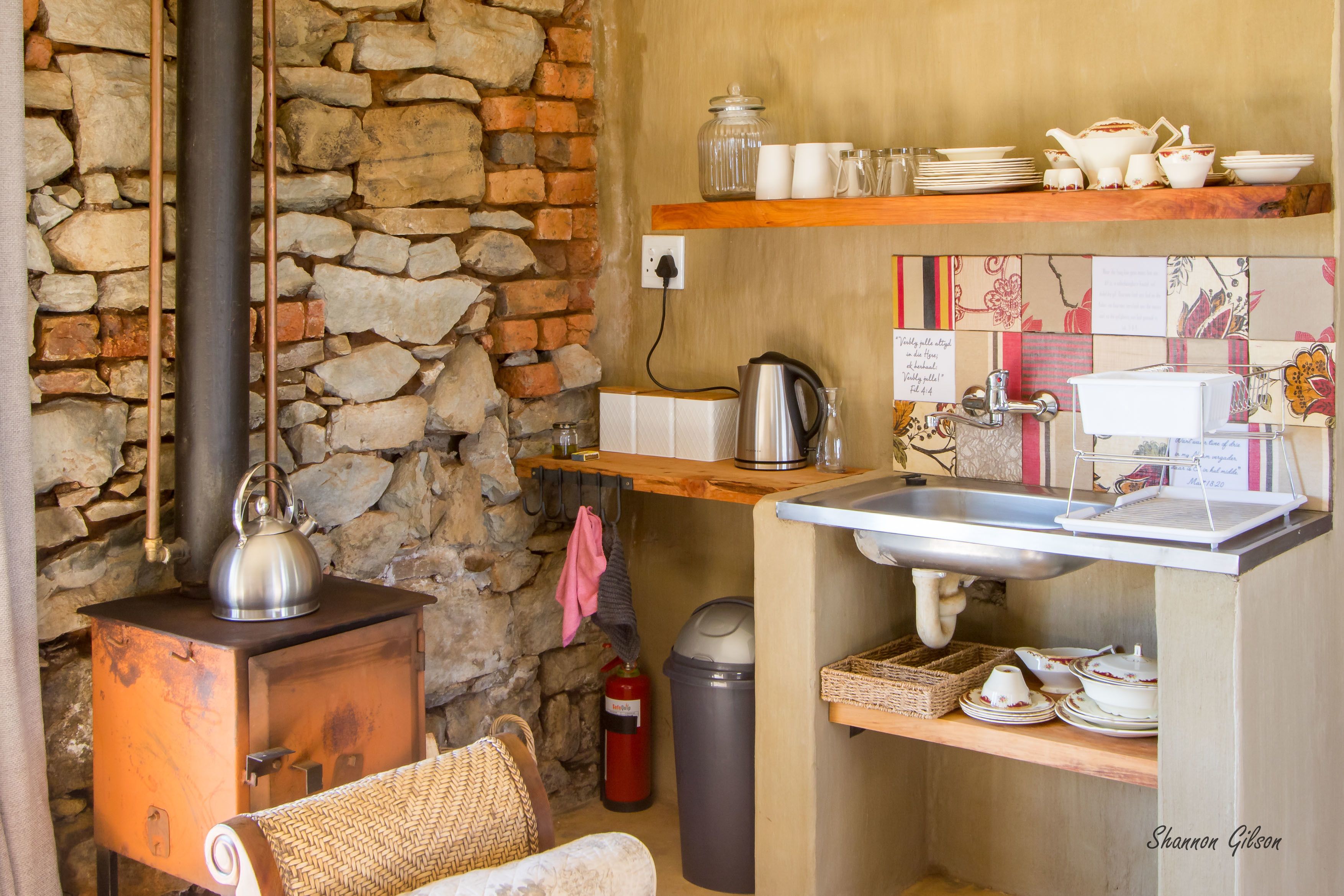 A rustic kitchen with a stone wall, a wooden stove, a sink, and shelves with dishes and a kettle.