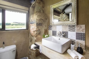 A bathroom with a stone wall, a white sink, a mirror, and a window with a view of the countryside.