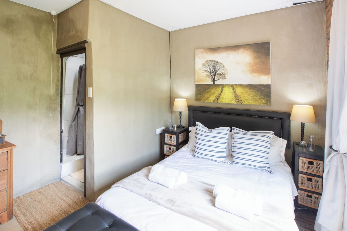 A neatly made bed with white linens and striped pillows, a headboard, bedside tables with lamps, a painting of a tree on the wall, and a wooden bench at the foot of the bed.
