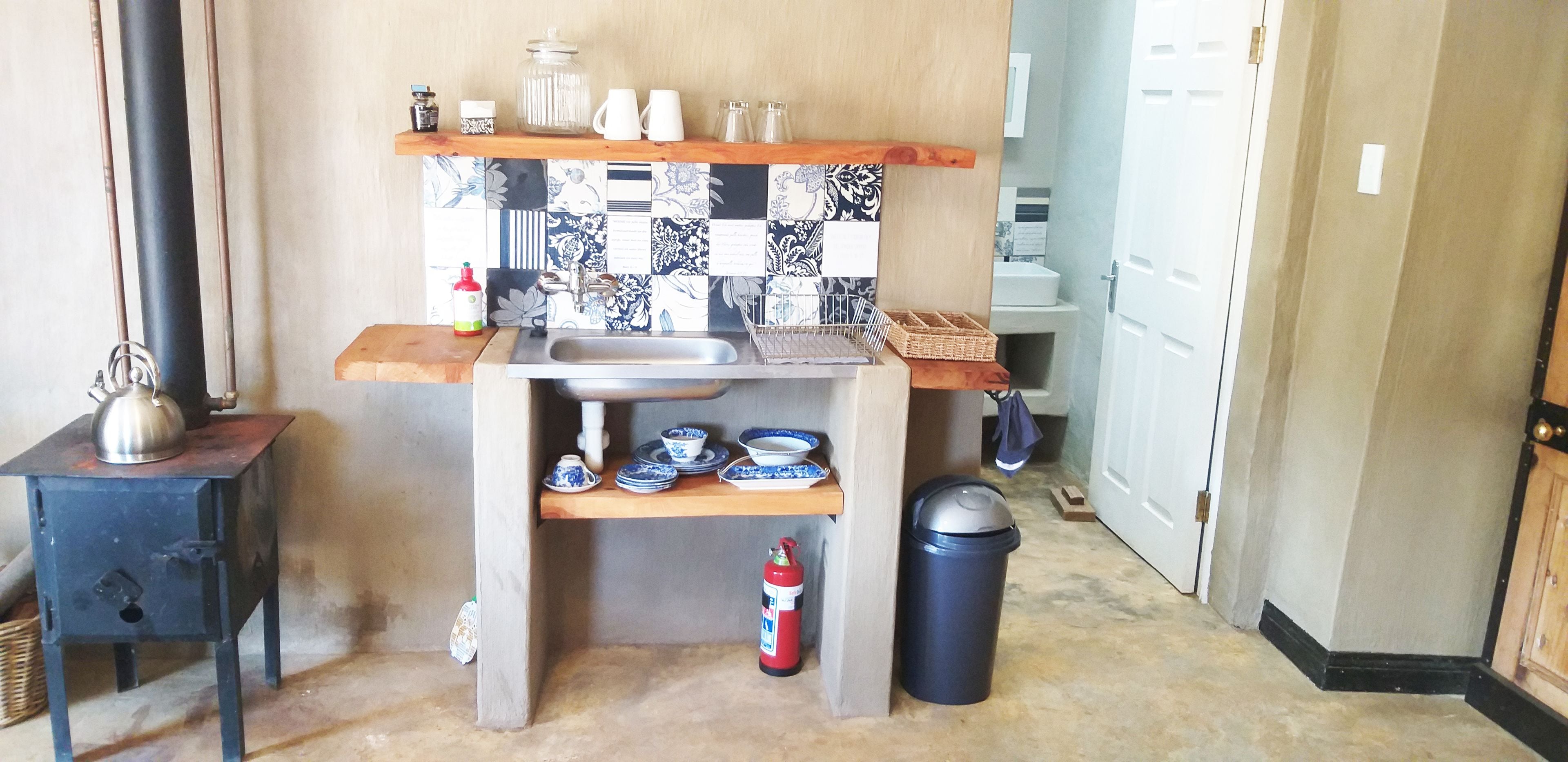 Una cocina compacta con un lavabo, estantes con vajilla, un hervidor y un cubo de basura.