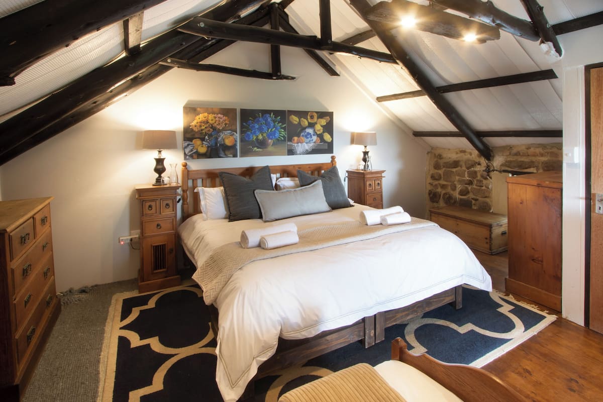 A bedroom with a large bed, wooden headboard, and a stone wall. There are two bedside tables with lamps, a dresser, and a bench.