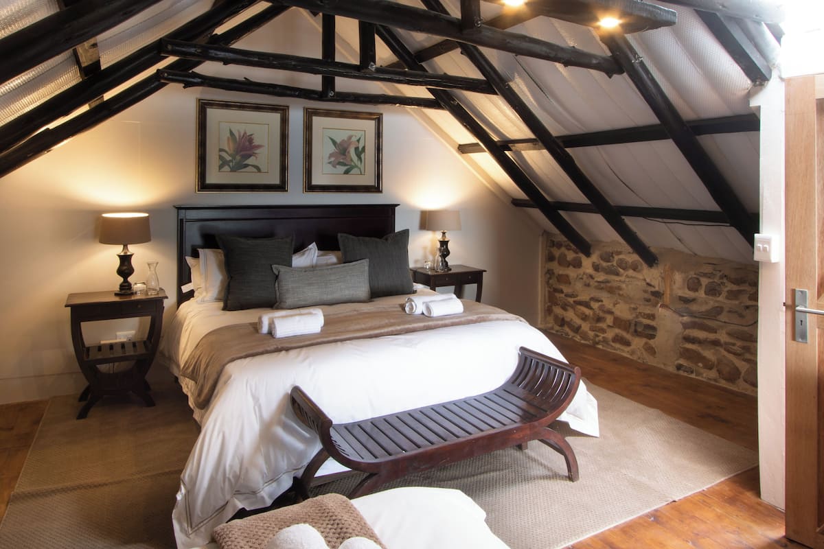 A bedroom with a large bed, two bedside tables with lamps, a wooden bench, and a stone wall.