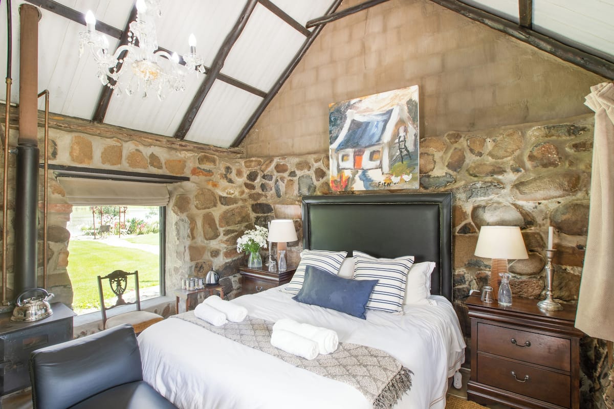 A bedroom with a stone wall, a bed with white bedding, a chandelier, and a painting above the bed.