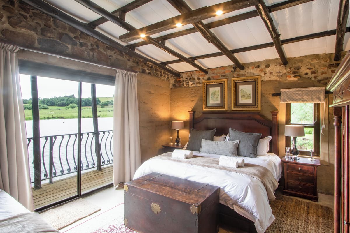 A bedroom with a large bed, a wooden headboard, two bedside tables with lamps, a nightstand, and a view of a green landscape through a balcony.