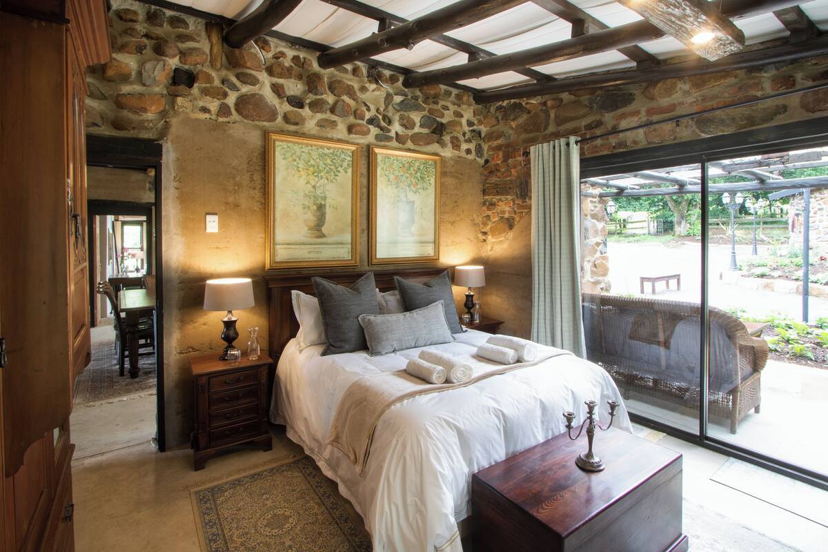A bedroom with stone walls, a large bed with white linens, two bedside lamps, and a view of the outdoors.