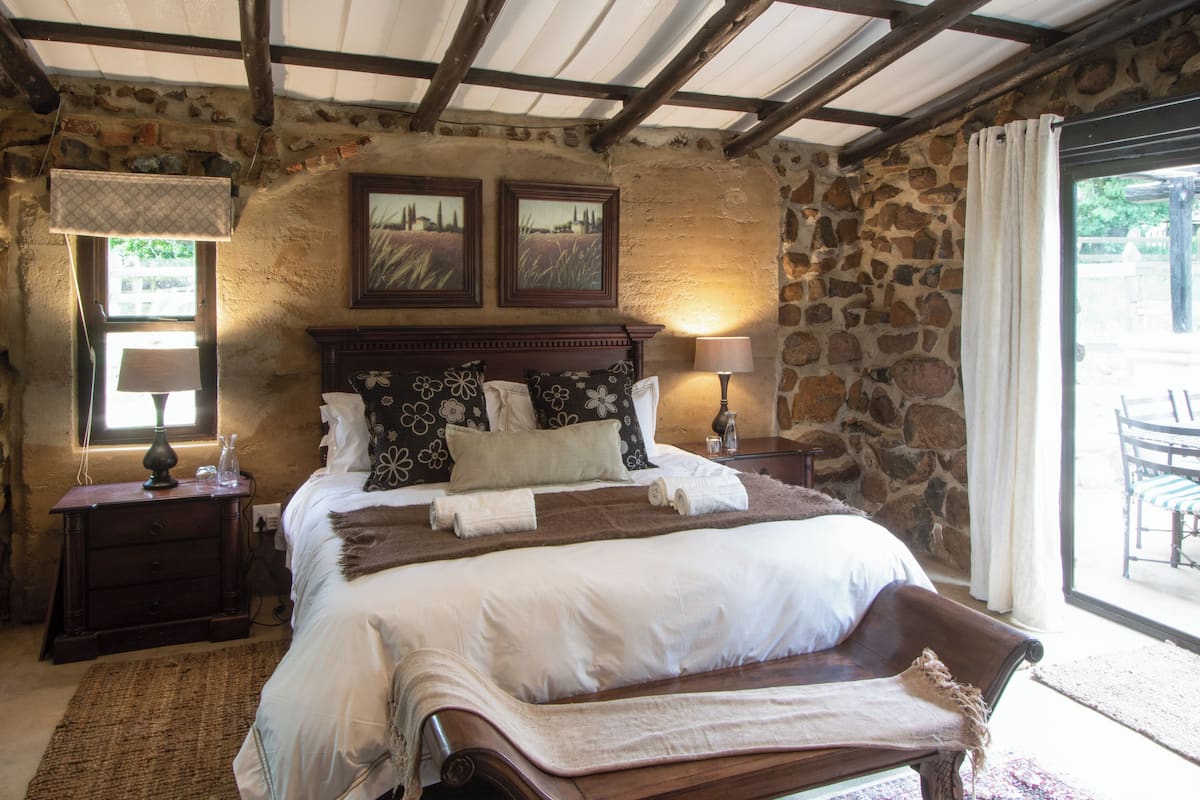 A bedroom with a stone wall, wooden bed frame, two bedside lamps, and a view of the outdoors.