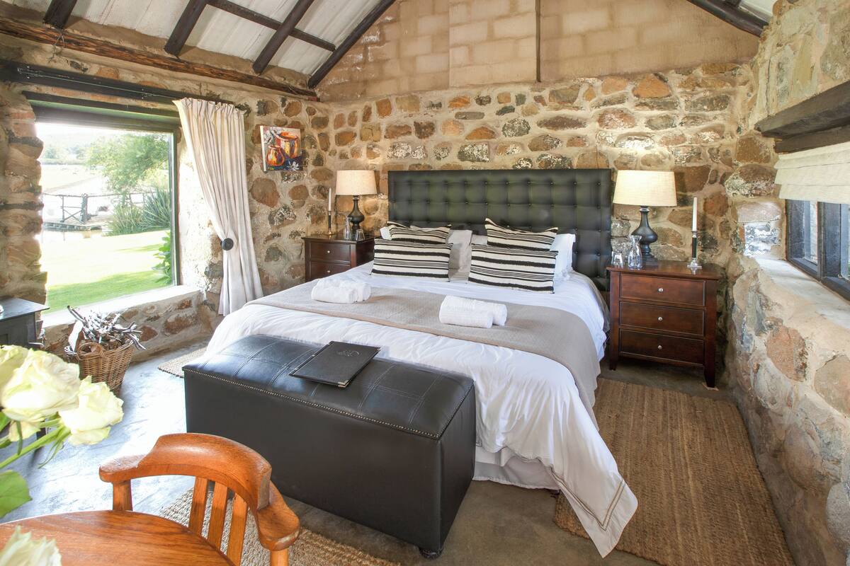 A bedroom with a stone wall, a bed with striped pillows, a wooden chair, and a nightstand with lamps.