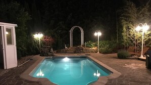 Piscina con baldosas de piedra, rodeada por un jardín con un arco de piedra e iluminación.