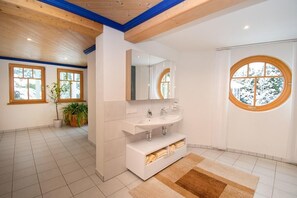A modern bathroom with a white sink, mirror, and built-in storage. There are potted plants and a large window with a view of trees. - Villa Salza (Mitterberg-Sankt Martin)
