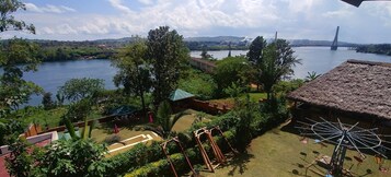 Vista junto al río con una cabaña de techo de paja, un columpio y un puente al fondo.