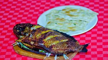 A grilled fish on a wooden board with a plate of flatbreads in the background.