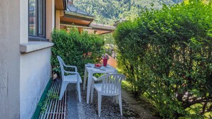 Un patio con due sedie bianche in plastica e un tavolo, immerso nel verde e con vista su una montagna.