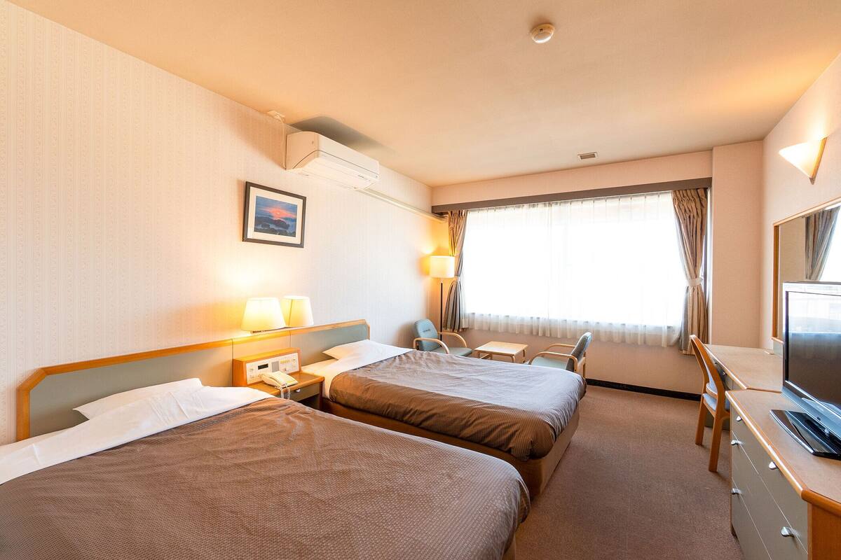 Ein Hotelzimmer mit zwei Betten, einem Schreibtisch, einem Sessel, einem Fernseher und einem Fenster mit Vorhängen.
