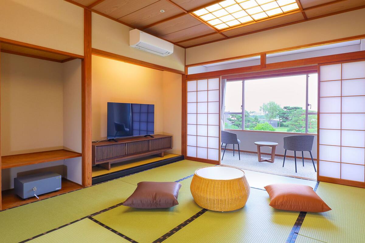 Ein traditioneller japanischer Raum mit Tatami-Böden, einem Fernseher auf einem niedrigen Holzständer, einem Tatami-Tisch mit Kissen und einer Schiebetür, die auf einen Balkon mit Blick auf Grün führt.