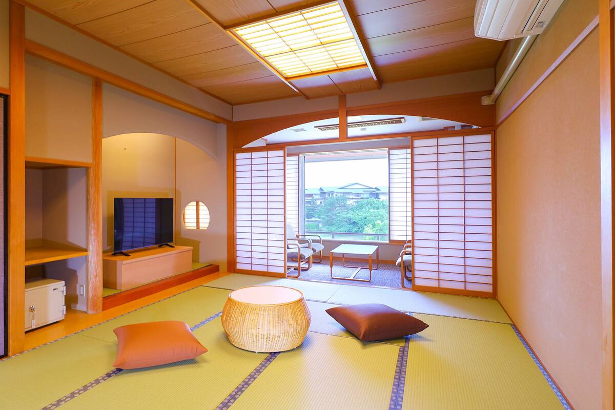 Ein traditioneller japanischer Raum mit Tatami-Böden, einem Fernseher, einem niedrigen Tisch und Blick auf einen Garten durch einen Shoji-Bildschirm.