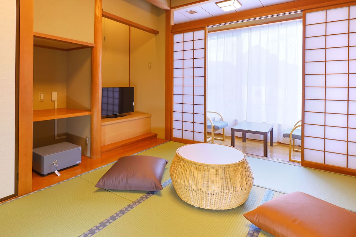 Ein traditioneller japanischer Raum mit Tatami-Bodenbelag, einem niedrigen Tisch mit Kissen, einem Fernseher auf einem Regal und Schiebetüren mit Rasteroptik.