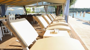 A row of white lounge chairs under a yellow canopy, with a wooden deck and a view of the sea. - Scheria Hotel (Corfu)