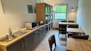 A compact kitchen with a microwave, sink, and cabinets, and a dining area with wooden tables and chairs. - T3 Budget Zwickau (Zwickau)