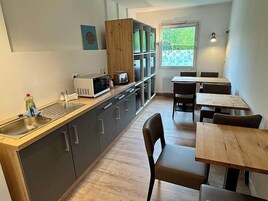 Cocina compacta con microondas, lavamanos y armarios, junto a un comedor con mesas y sillas de madera.
